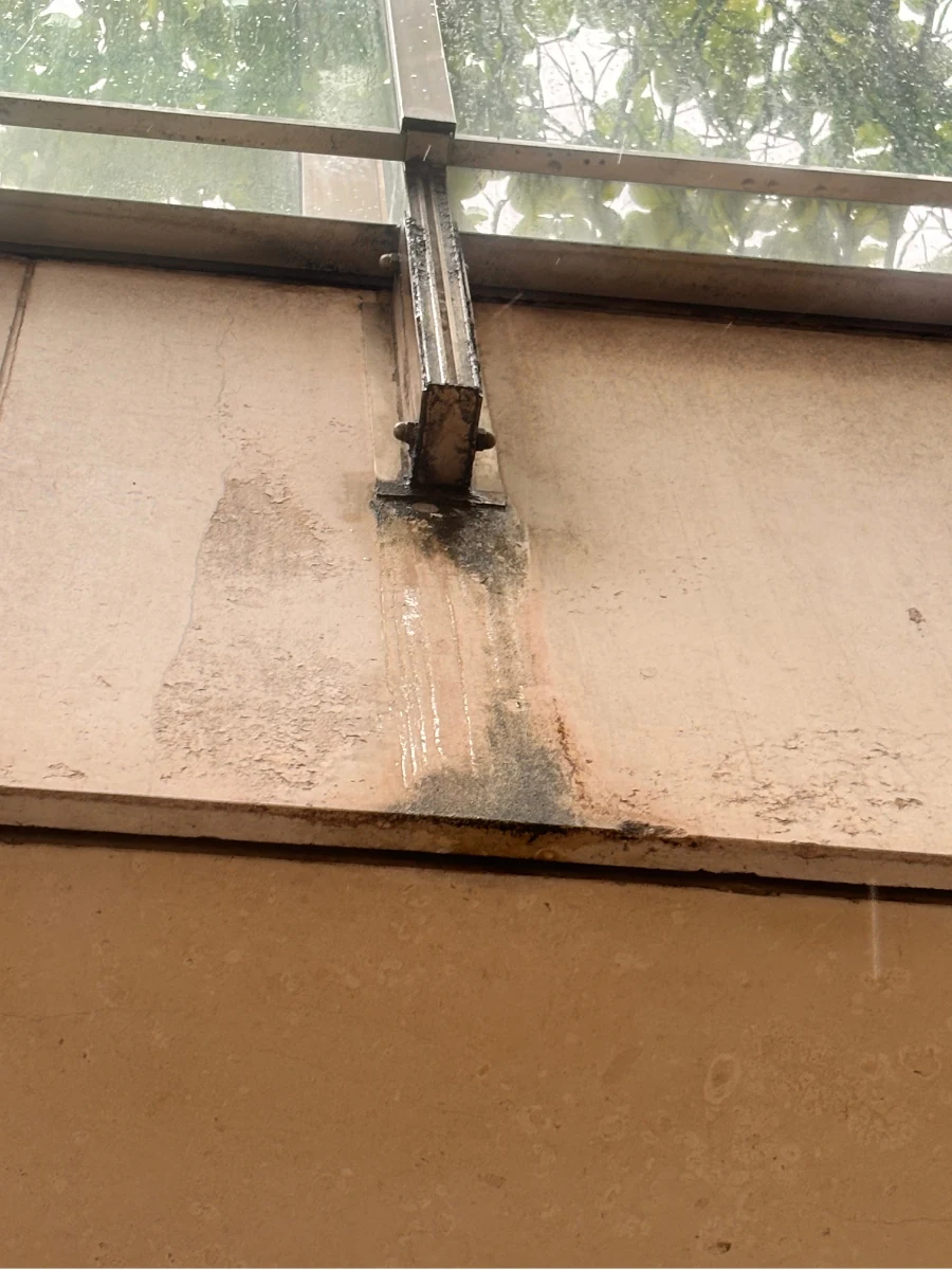 Base de guarda-corpo em aço inox com acúmulo de material orgânico escuro e marcas de escorrimento sobre a pedra.